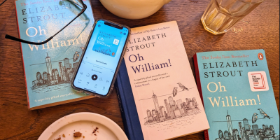 Photo of three physical copies of the book, "Oh William!" on a table, alongside a phone showing a narrated version. There is also a pair of reading glasses, a teapot, an empty glass, and an empty plate (that looks very much as though it formerly contained a most excellent chocolate cake), signifying the end of a very enjoyable NTU Psychology Book Club session.